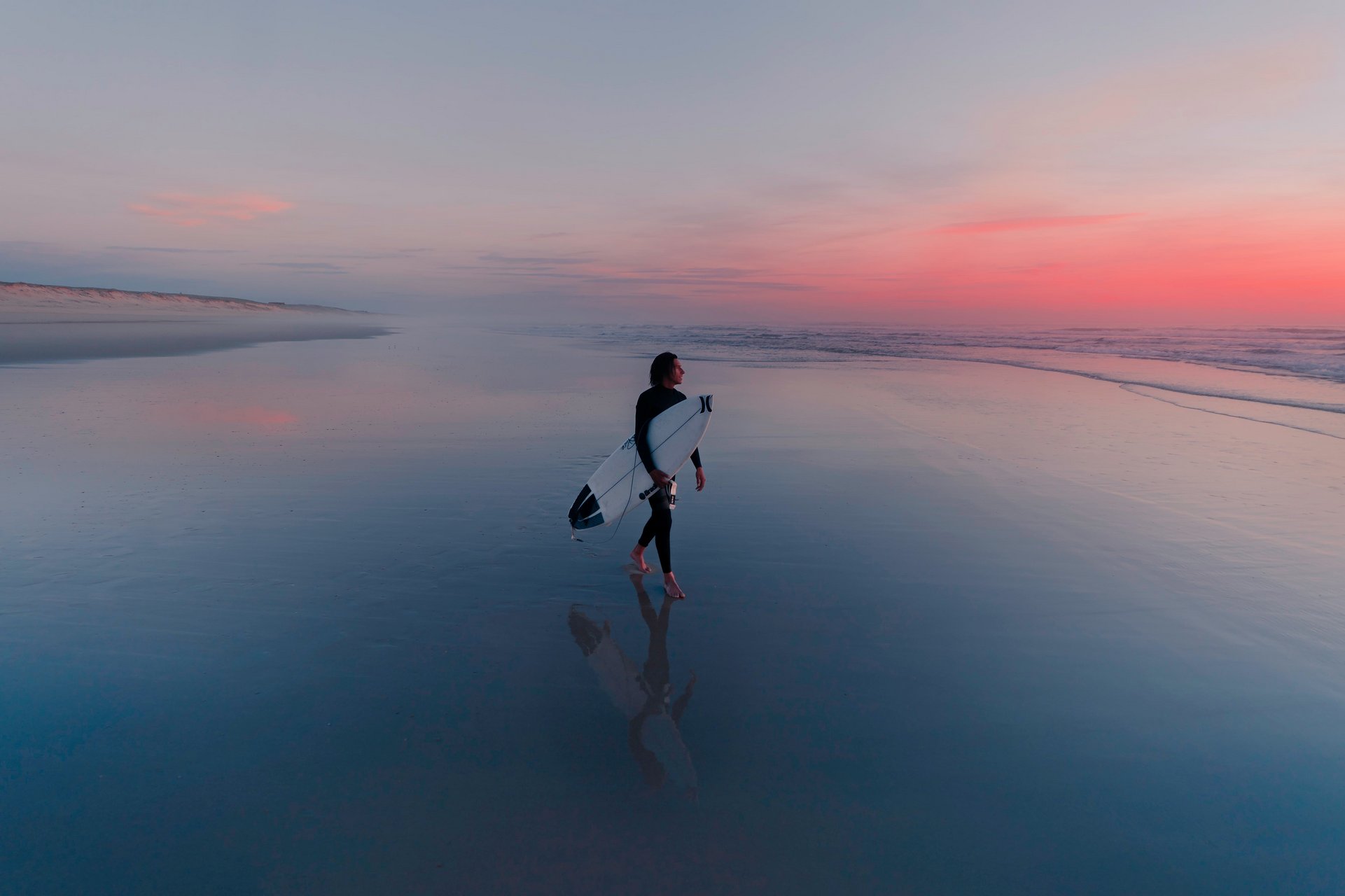 Ein Surfer im Neoprenanzug trägt bei Sonnenuntergang sein Surfbrett